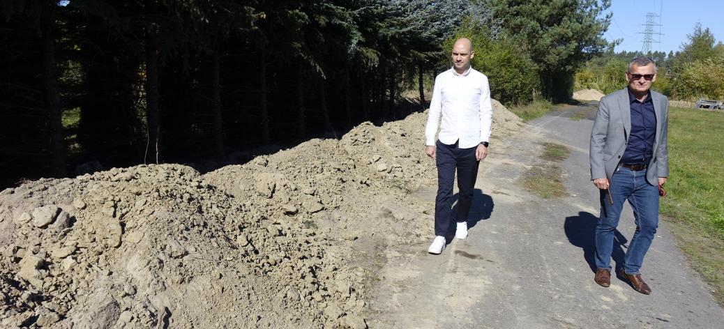  Burmistrz (po prawej) i Wiceburmistrz Drzewicy idą asfaltową drogą wzdłuż wykopu ziemnego i pasa iglastych drzew.