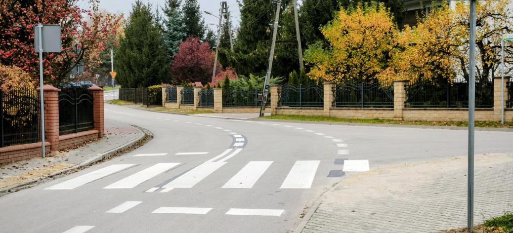 Przejście dla pieszych na łuku asfaltowej drogi z pylonami i znakiem D-6 w otoczeniu jesiennej zieleni i domów jednorodzinnych.