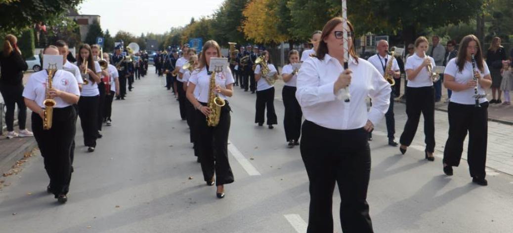 Przemarsz Młodzieżowej Orkiestry Dętej z Drzewicy ulicą miasta. Na czele idzie kapelmistrzyni w białej koszuli i czarnych spodniach, trzymając buławę. Za nią maszerują młodzi muzycy grający na instrumentach dętych, m.in. saksofonach i klarnetach.