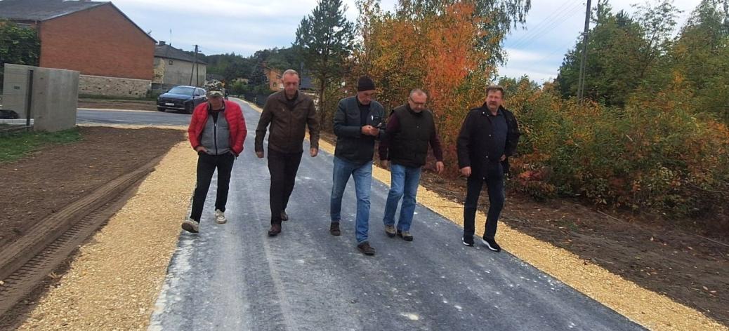 Pięciu mężczyzn idących nowo wybudowaną drogą asfaltową w Brzustowcu-Smugach w otoczeniu zabudowań i jesiennej zieleni. 