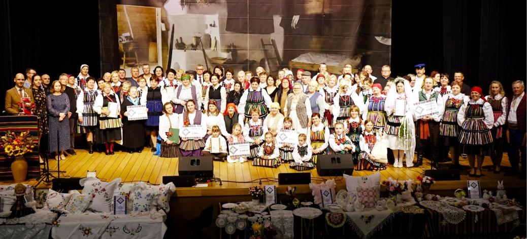 Zdjęcie grupowe uczestników Rewii na scenie sali widowiskowej RCK. W centrum osoby w tradycyjnych strojach ludowych, dzieci oraz goście. Na pierwszym planie stoły nakryte haftowanymi obrusami z wyrobami rękodzieła i poduszkami dekoracyjnymi. W tle dekoracja sceniczna z motywem dawnej chaty.