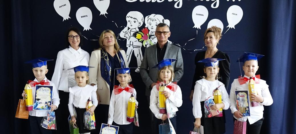  Sześcioro dzieci w biretach i strojach galowych pozujących do wspólnego zdjęcia z dyrekcją, kadrą pedagogiczną i gośćmi na tle dekoracji z napisem „Uroczyście Ślubujemy...”.