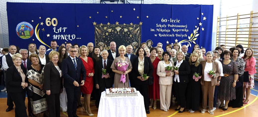 Pozowane zdjęcie grupowe kilkudziesięciorga uczestników jubileuszu 60-lecia SP w Radzicach Dużych. Przed grupą stoi stolik z uroczystym tortem, a w tle znajduje się niebieska ścianka dekoracyjna. 