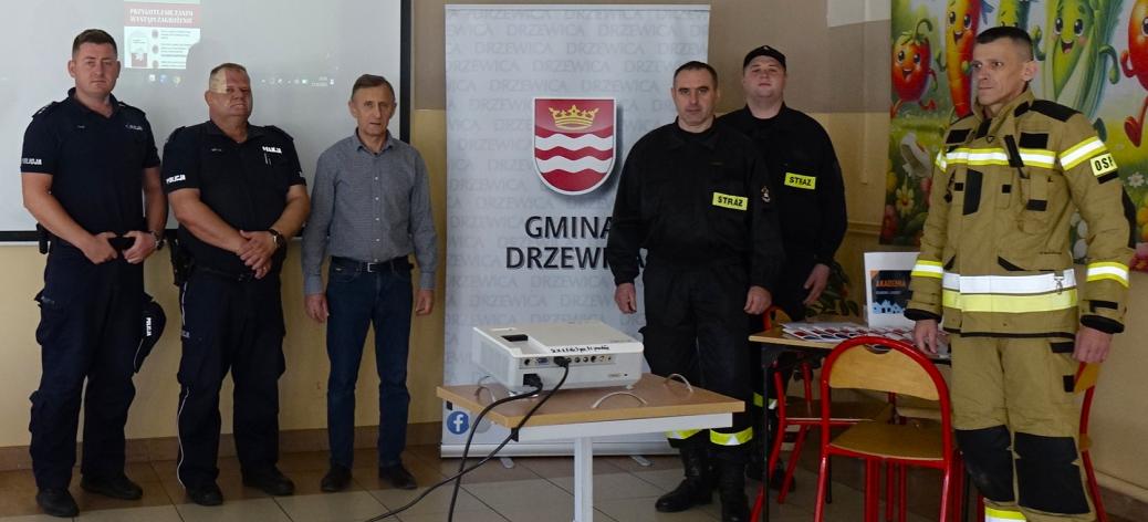 Grupa sześciu mężczyzn (policjanci, strażacy, cywil) stoi w rzędzie w sali prelekcyjnej między dwoma banerami reklamowymi a ekranem projekcyjnym.