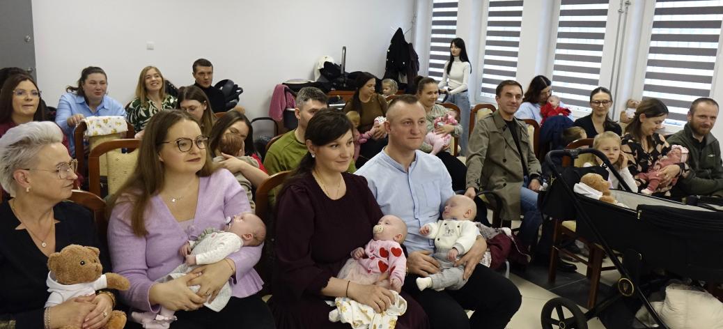 Grupowe zdjęcie uczestników spotkania w sali USC. Rodzice z niemowlętami na rękach oraz małe dzieci siedzą w krzesłach i prezentują otrzymane maskotki misiów oraz pamiątkowe teczki. 