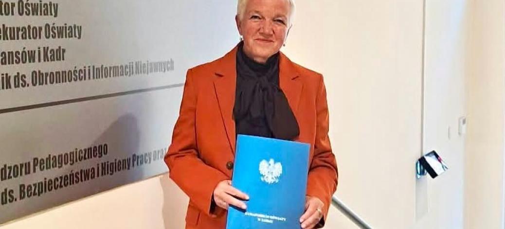 Bożena Ramus w rudym garniturze trzyma niebieską teczkę z godłem Polski we wnętrzu budynku Kuratorium Oświaty. 