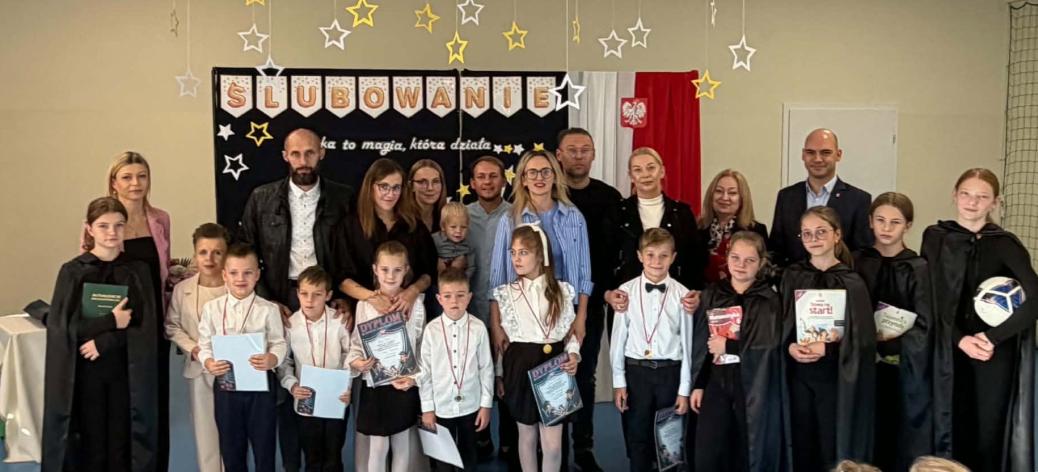 Szkolna uroczystość ślubowania. Zdjęcie grupowe pierwszoklasistów, ich rodziców, starszych uczniów oraz nauczycieli i gości na tle dekoracji z napisem „ŚLUBOWANIE”. 