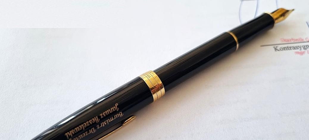  Eleganckie, czarne wieczne pióro ze złotą stalówką i złotymi wykończeniami, leżące na białym dokumencie papierowym. Na korpusie pióra znajduje się grawerowany napis w kolorze złotym: „Burmistrz Drzewicy Janusz Reszelewski”. W tle widoczny jest fragment podpisu i pieczątki na dokumencie.