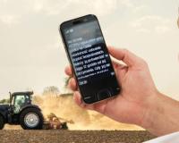 Na pierwszym planie dłoń a w niej telefon z tekstem wiadomości, w tle traktor na polu
