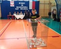 Wybory do MRM w roku 2024. W sali gimnastycznej kartę do głosowania wrzuca do urny wyborczej uczeń SP w Brzustowcu. W tle szkolna komisja wyborcza