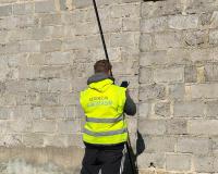Mężczyzna w kamizelce odblaskowej wykonuje pomiar przy ścianie budynku za pomocą sprzętu geodezyjnego.