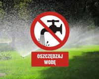 Znak zakazu z kranem oraz napis „OSZCZĘDZAJ WODĘ” na tle zraszanego trawnika.