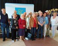 Grupowe zdjęcie kilkunastu seniorów, stojących blisko siebie w pomieszczeniu i pozujących do fotografii. Grupa składa się z kobiet i mężczyzn ubranych w stroje codzienne i wyjściowe. W tle czarna kotara, złote balony oraz jasna tablica z niebiesko-żółtym herbem przedstawiającym splecione litery. Po prawej stronie baner typu roll-up.