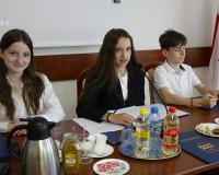 Troje młodych radnych przy stole prezydialnym podczas obrad w Drzewicy. W środku Przewodnicząca Młodzieżowej Rady Miejskiej wypowiada się do mikrofonu; na stole widoczne są dokumenty, napoje oraz oficjalne flagi w tle.