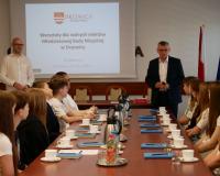 Na zdjęciu spotkanie warsztatowe: grupa młodzieży siedzi przy stole konferencyjnym, po prawej stronie stołu Burmistrza Drzewicy zabierający głos, po lewej Zastępca Burmistrza. Na ekranie wyświetlony jest slajd dotyczący warsztatów dla Młodzieżowej Rady Miejskiej w Drzewicy