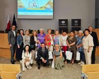 W nowoczesnej sali konferencyjnej stoi grupa kilkunastu osób, w większości kobiet, trzymających białe torby upominkowe. Po lewej stoi Wiceburmistrz Drzewicy, a z przodu kucają dwie kobiety i mężczyzna. W tle drewniane podwyższenie, flagi Polski i UE, dwa czarne banery z napisami „Łódzki Urząd Wojewódzki w Łodzi” oraz „Wojewoda Łódzki”, a także duży ekran projekcyjny wyświetlający slajd z napisem „Świadomy Senior Bezpieczny Senior”.