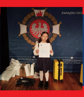 Trzymająca dyplom dziewczynka stoi na tle loga Związku OSP RP. Po obydwu stronach dziecka nagrody rzeczowe
