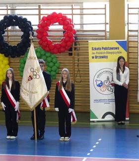 Oficjalne otwarcie gali w hali sportowej. Od lewej: trzyosobowy szkolny poczet sztandarowy na tle kół olimpijskich, uczennica Michalina Jedynak i Dyrektor SP w Drzewicy Agnieszka Bogatek