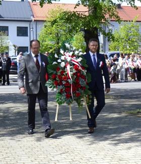 Dwaj pracownicy Urzędu Miejskiego w Drzewicy zmierzają pod Pomnik Pamięci, niosąc wieniec z białymi i czerwonymi kwiatami