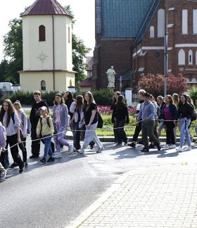 Grupa pielgrzymów wychodzi na trasę. Maszeruje ulicą na placu Wolności w Drzewicy. W tle m.in. kościół i dzwonnica.