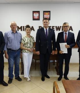 3.	Wspólne zdjęcie. W sali konferencyjnej Urzędu Miejskiego w Drzewicy, na tle ściany z napisem Gmina Drzewica, herbem Drzewicy oraz godłem Rzeczpospolitej Polskiej stoją: (od lewej): Sołtys Miejscowości Świerczyna Karol Siedlecki, Sołtys Miejscowości Idzikowice Roman Mastalerek, Skarbnik Gminy Drzewica Irena Jóźwicka, Wicemarszałek Województwa Łódzkiego Artur Bagieński, Burmistrz Drzewicy Janusz Reszelewski, Zastępca Burmistrza Drzewicy Dominik Niemirski. 