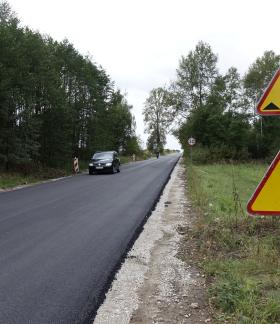 Widok remontowanej drogi powiatowej z nową warstwą ścieralną. Po drodze jedzie auto. Z prawej znaki informujące o robotach drogowych