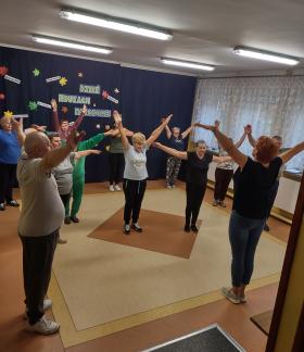 Grupa seniorów na zajęciach fitness w SP w Idzikowicach. Na pierwszym planie instruktor zajęć Alina Szymańska, na planie dalszym uczestnicy zajęć patrzący na instruktora wykonują ćwiczenia