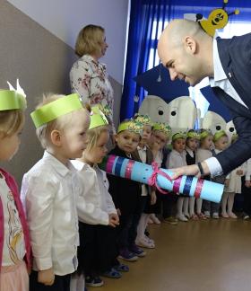 Zastępca Burmistrza Drzewicy Dominik Niemirski pasuje na przedszkolaków dzieci z grupy „Skrzaty”. Pochylając się, wielki kolorowy ołówek kładzie na ramieniu dziewczynki.