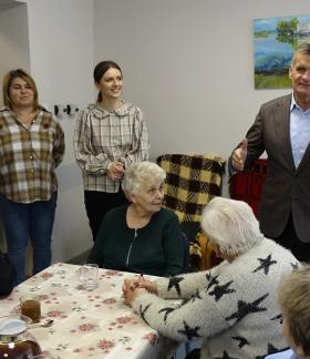 Burmistrz Drzewicy Janusz Reszelewski składa seniorom życzenia. Obok po lewej stoją: Kierownik DDS+ Paulina Klata, pracownica i uczestnik Seniora+. Poniżej przy stole siedzi czworo seniorów.