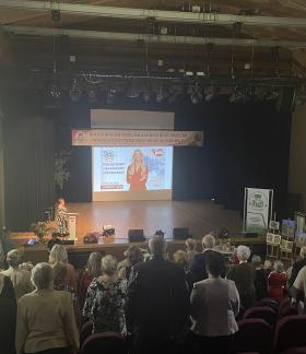 Inauguracja roku akademickiego UTW w Drzewicy. Zdjęcie z góry publiczności. Zgromadzeni stoją podczas odśpiewania hymnu „Gaudeamus igitur”. W tle scena.