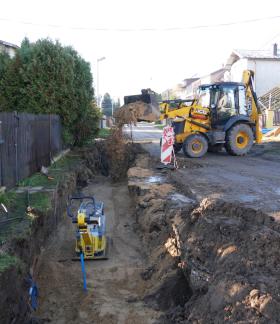 Przebudowa sieci wodociągowej na Os. Słonecznym w Drzewicy. Na zdjęciu wykop, w środku którego znajduje się zagęszczarka. W tle koparko-ładowarka zasypuje wykop drobnym piaskiem. Wokoło zabudowania przy ulicy. 