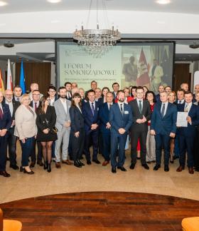 Wspólne zdjęcie kilkudziesięcioosobowowej grupy uczestników Forum stojących w przestronnej sali konferencyjnej. W tle ekran, flagi, rull-upy, na górze żyrandol