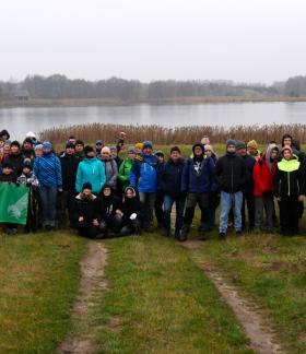 Wspólne zdjęcie kilkudziesięcioosobowej grupy piechurów stojących na trawie na tle Zalewu Drzewickiego