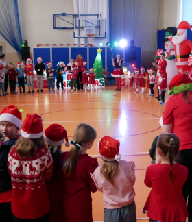 Mikołajki w SP w Brzustowcu. Grupa uczniów wraz z pracownikami szkoły zgromadzona w okręgu w sali gimnastycznej. Wokoło dmuchane dekoracje świąteczne – bałwany, mikołaje, renifery, żołnierzyki.