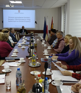 Spotkanie komitetu w Sali konferencyjnej Urzędu Miejskiego w Drzewicy. Na pierwszym planie przy stole głównym członkowie komitetu. W tle przy stole prezydialnym Zastępca Burmistrza Drzewicy Dominik Niemirski. Na ścianie za jego plecami na ekranie prezentacja multimedialna. 