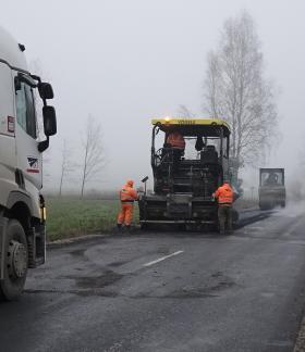 Trwają prace związane z modernizacją drogi powiatowej Krzczonów-Jelnia. Na drodze stoją: na pierwszym planie samochód ciężarowy, dalej rozściełacz do asfaltu a przy nim pracujący drogowcy. W tle dwa walce na asfalcie. 