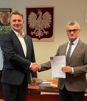 Członek Zarządu Woj. Łódzkiego Artur Bagieński i Burmistrz Drzewicy Janusz Reszelewski podają sobie dłonie. Burmistrz trzyma aneks. Po lewej flaga Polski, w tle godła Polski i Woj. Łódzkiego