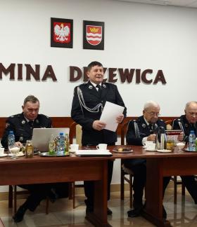 Sześciu strażaków za stołem prezydialnym zebrania w sali konferencyjnej UM w Drzewicy. Pięciu siedzi, a stojąc głos zabiera Gminny Komendant ZOSP RP w Drzewicy
