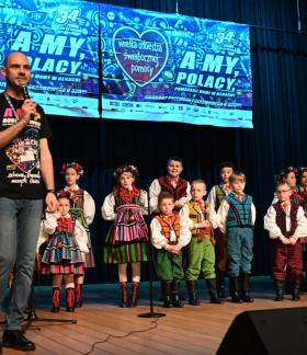 Na scenie w sali widowiskowej RCK z mikrofonem stoi szef drzewickiego sztabu WOŚP Dominik Niemirski. Za nim ubrani w stroje opoczyńskie ,,Mali Drzewiczanie'