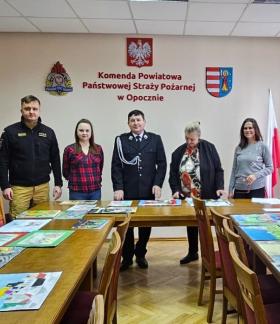 W sali konferencyjnej KP PSP w Opocznie za stołami, na których rozłożone są prace plastyczne, stoi pięć osób - dwóch strażaków i trzy panie.  W środku Gminny Komendant ZOSP RP w Drzewicy Marian Kalużny