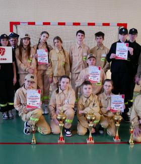 Młodzieżowe Drużyny Pożarnicze z OSP Domaszno wraz z opiekunami prezentują zdobyte trofea. Po lewej stoi Burmistrz Drzewicy.  W tle bramka do piłki ręcznej.