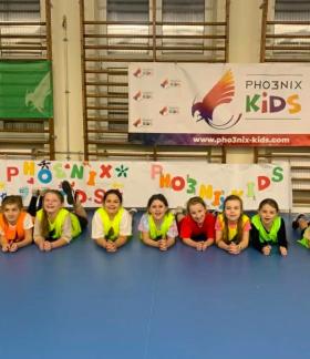 W hali sportowej dziesięcioro dzieci leży na parkiecie przodem do fotografa. W tle drabinki i banery Pho3nix Kids.