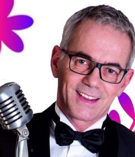  Uśmiechnięty Robert Janowki w marynarce i z muszką - w ujęciu od ramion - trzyma mikrofon retro. W tle kolorowe roślinne grafiki