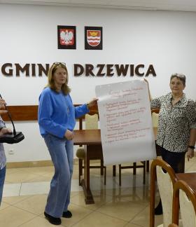 W sali konferencyjnej UM na tle napisu Gmina Drzewica stoi mówiący do mikrofonu Zastępca Burmistrza Drzewicy oraz troje radnych Miejskiej Rady Seniorów. Dwie radne trzymają zapisany arkusz papieru