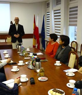  Zastępca Burmistrza Drzewicy Dominik Niemirski prowadzi szkolenie dla pań z KGW siedzących przy stole konferencyjnym. W  tle ekran z napisem ,,O obowiązkach KGW w księgowości'' oraz flagi