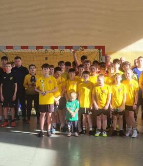 Grupowe zdjęcie kilku młodzieżowych drużyn z organizatorami turnieju na tle bramki w hali sportowej SP w Drzewicy