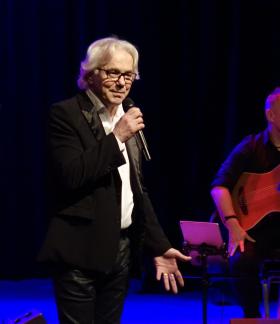 Na scenie z mikrofonem stoi Robert Janowski. Po prawej stronie w tle siedzi gitarzysta.