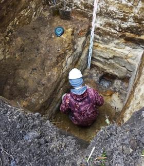 W głębokim wykopie, kucając pracuje archeolog z kaskiem na głowie (widok na jego plecy). Powyżej miarka głębokości.