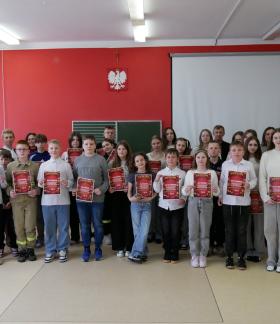 Na zdjęciu widoczna jest grupa uczniów wraz z nauczycielami stojących w sali lekcyjnej. Dzieci ustawione są w kilku rzędach i trzymają w rękach dyplomy. Za nimi znajduje się tablica szkolna oraz rozwinięty ekran projekcyjny. Ściana w tle jest czerwona, a na niej umieszczone są krzyż oraz godło Polski (biały orzeł na czerwonym tle). Wszyscy pozują do wspólnego zdjęcia.