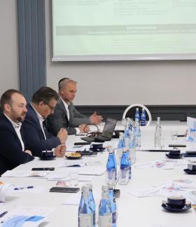 Na zdjęciu spotkanie odbywające się w sali konferencyjnej. Przy długim stole siedzi kilkanaście osób, przeglądają dokumenty, notują oraz słuchają osoby mówiącej.  W tle widać ekran projekcyjny z wyświetloną prezentacją.